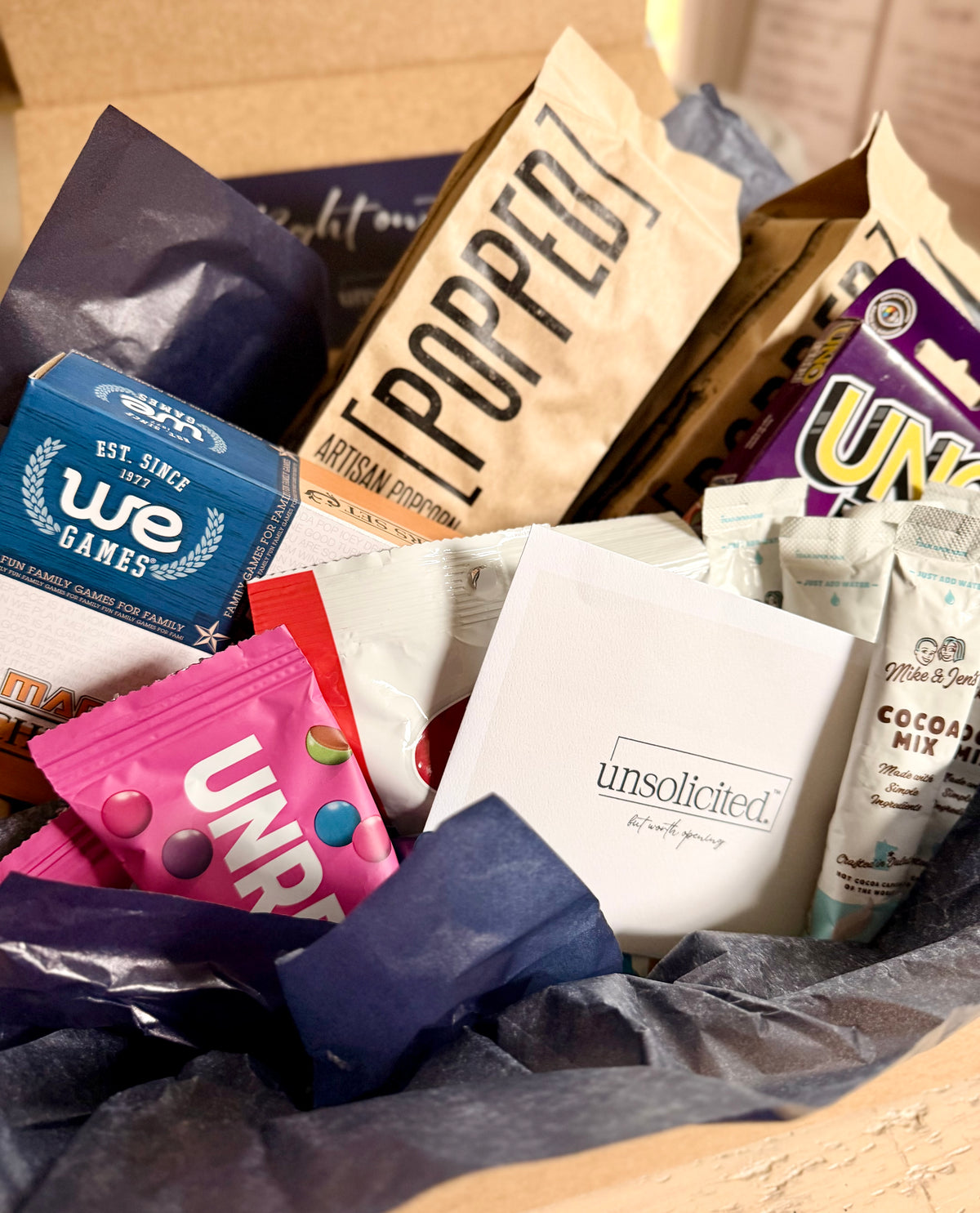 Open unsolicited. care package with a letter, popcorn, candy, cocoa mix, and games in navy tissue.
