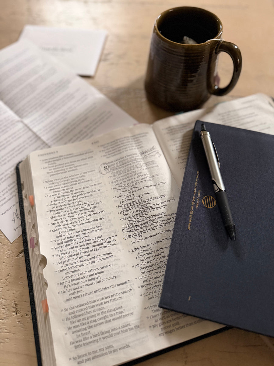 Open Bible beside a navy notebook, pen, and ceramic mug on a wooden table.